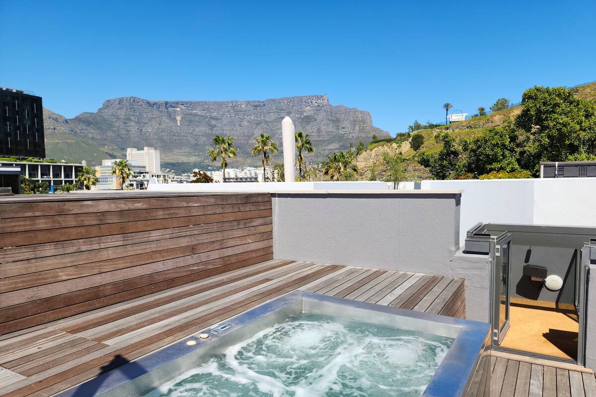 A rooftop with a hot tub, wooden deck, and a view of mountains and buildings.