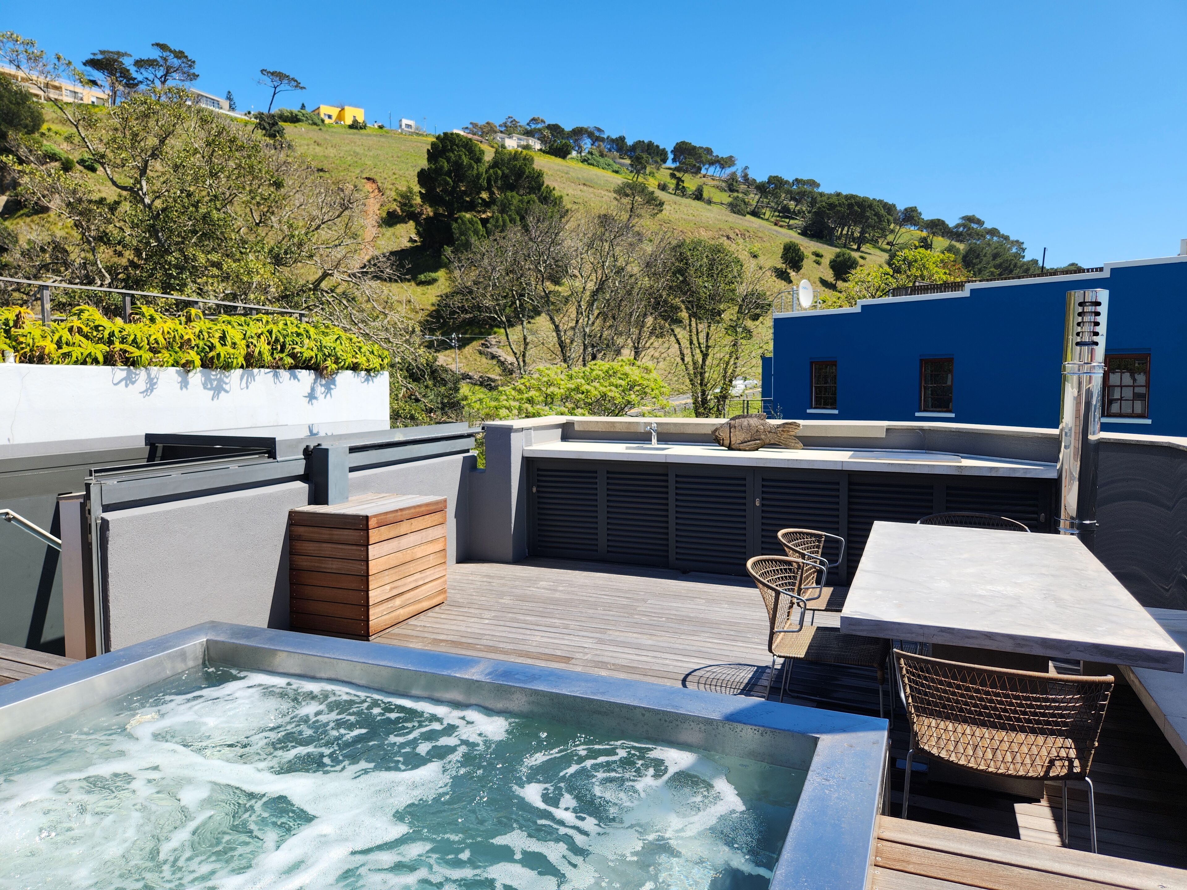 A rooftop terrace with a hot tub, seating area, and a view of a hillside with buildings.