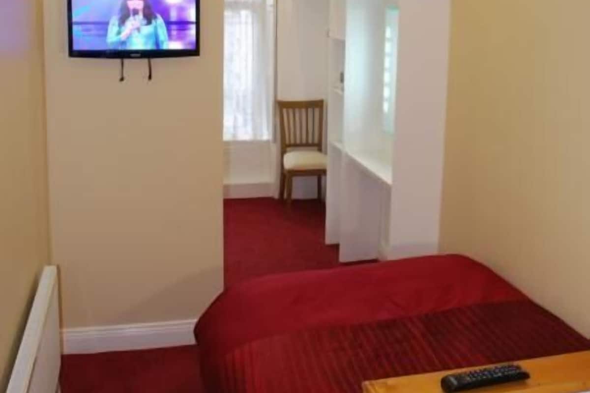 Ein Hotelzimmer mit an der Wand montiertem Fernseher, einem Bett mit roter Decke, einem Holzstuhl und einem weißen Kleiderschrank.