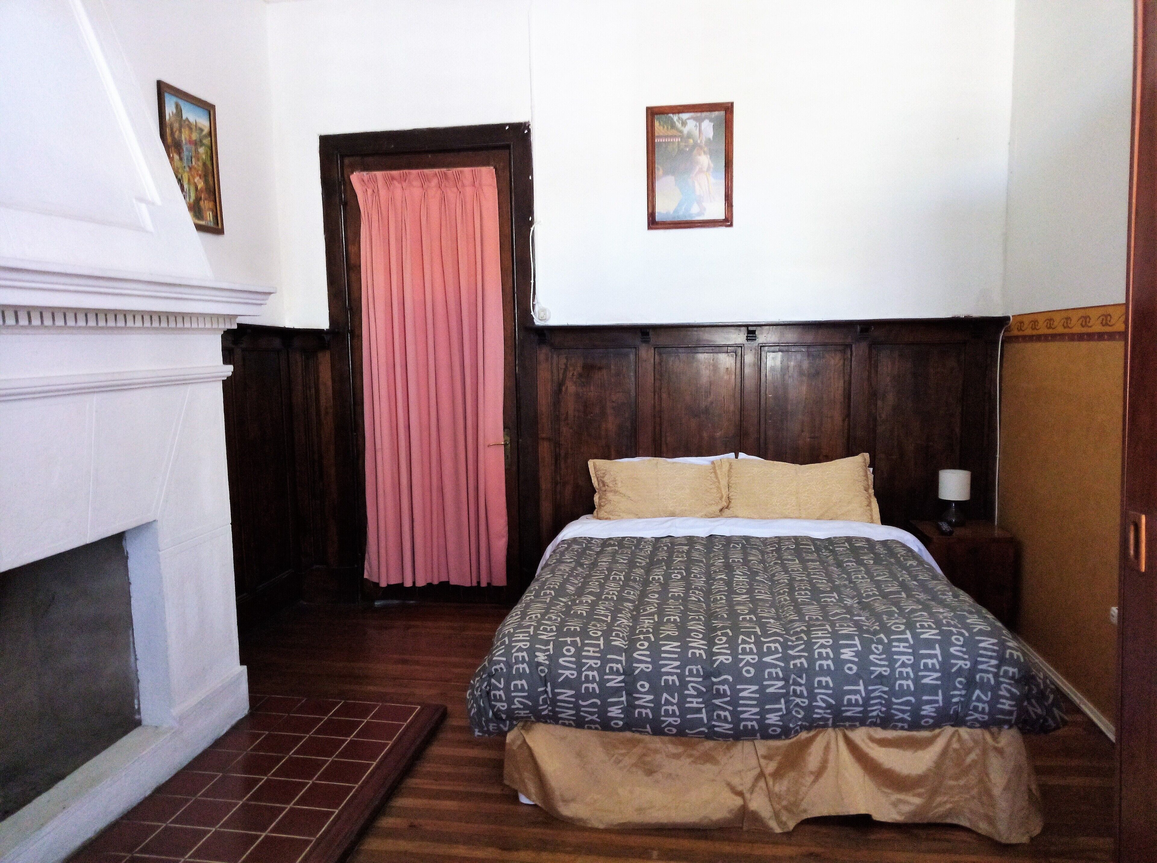 A bedroom with a wooden headboard, a bed with patterned bedding, a pink curtain, and a small framed picture on the wall.