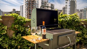 Een draagbare grill met een fles wijn en glazen op een houten tafel, omringd door groen, met hoge gebouwen op de achtergrond.