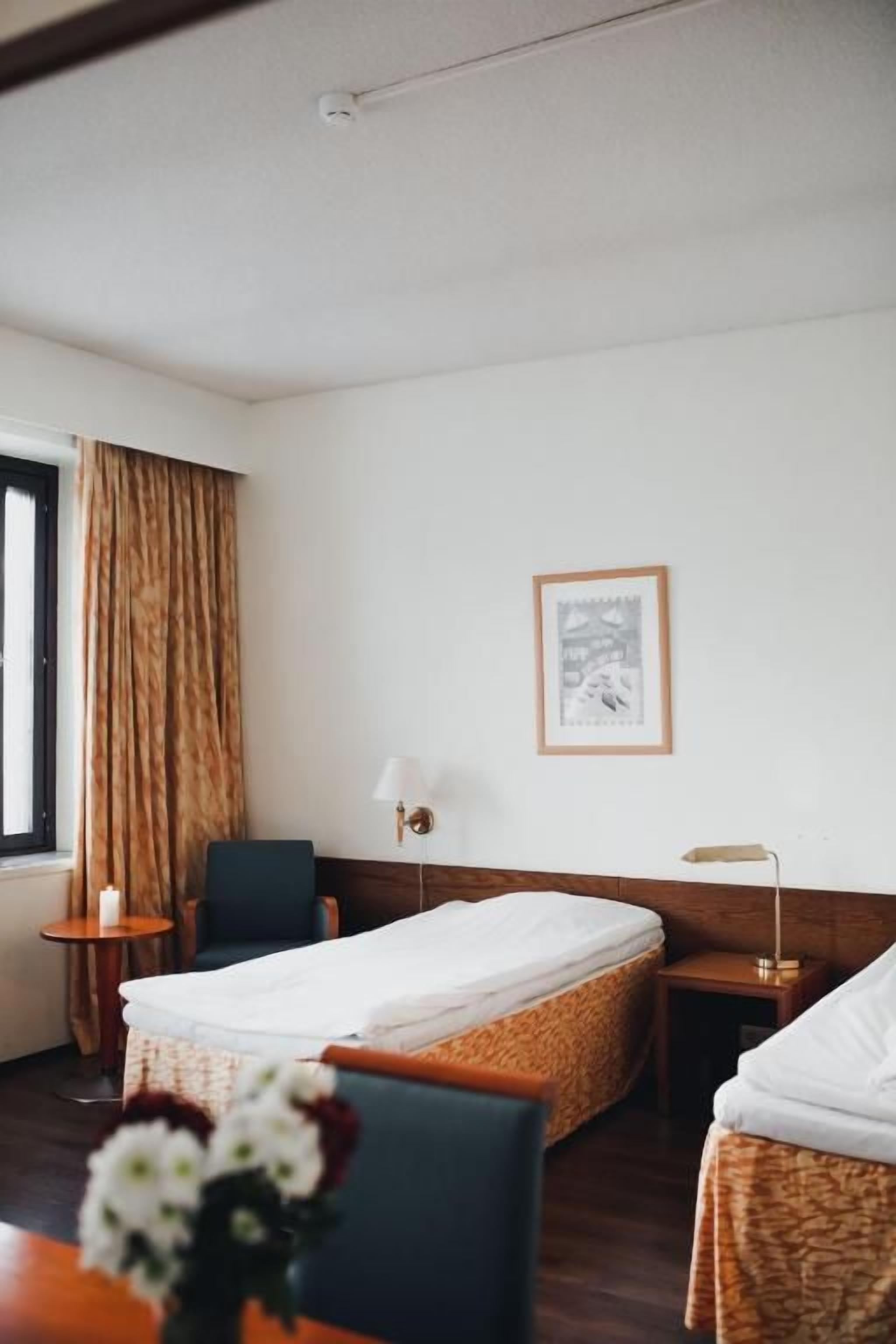 A hotel room with a bed, a chair, a small table, a lamp, and a framed picture on the wall.