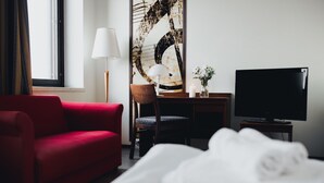 Habitación de hotel con sofá rojo, escritorio con lámpara, televisor y cama con sábanas blancas.