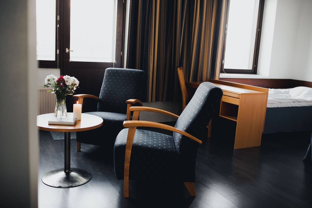 Hotellihuone, jossa on sänky, työpöytä, kaksi tuolia, pieni pöytä, jossa on maljakko täynnä kukkia, ja ikkuna, jossa on verhot.