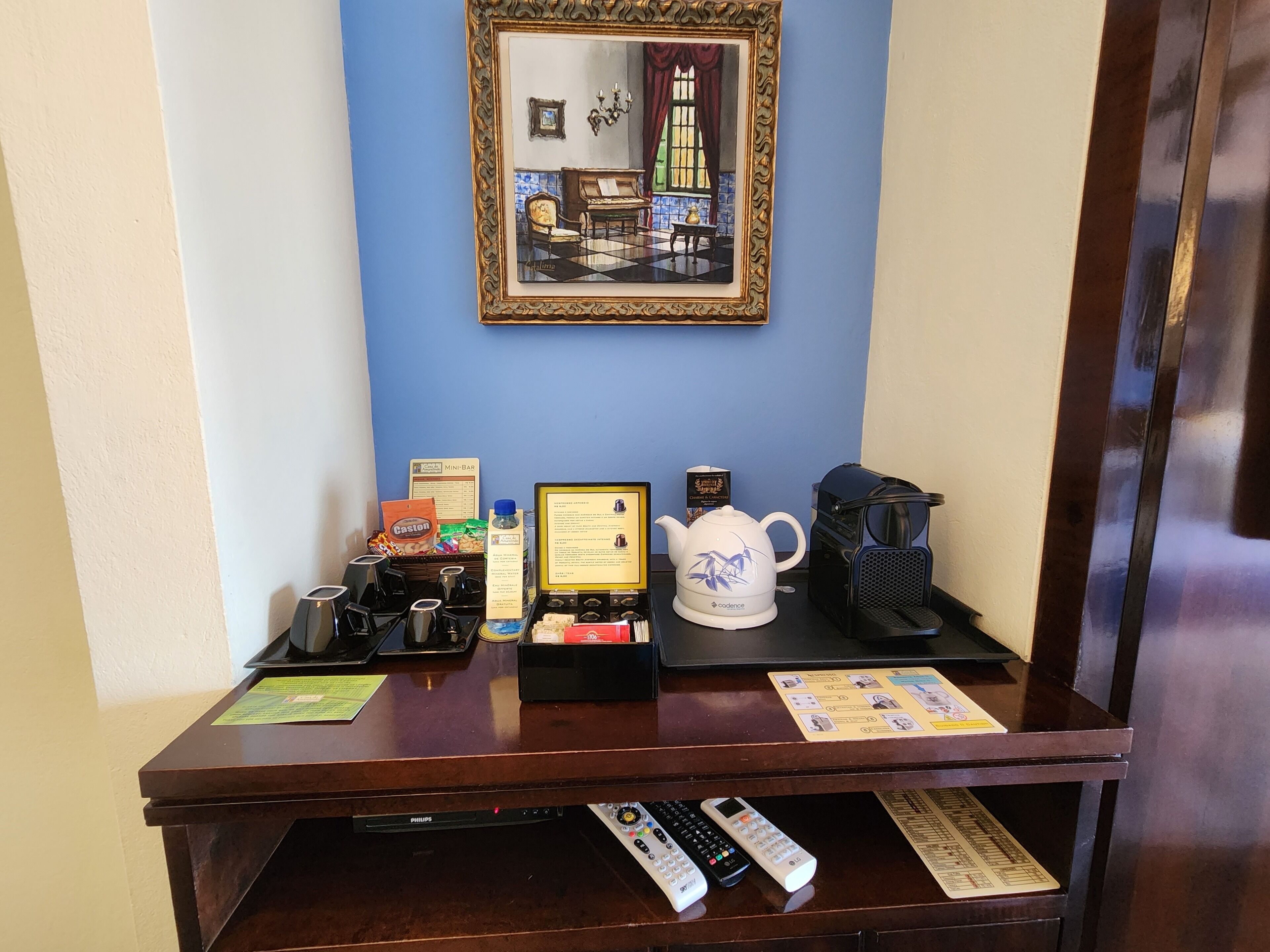 A reception desk with a framed picture, a coffee machine, a mini-bar, and a set of remote controls.
