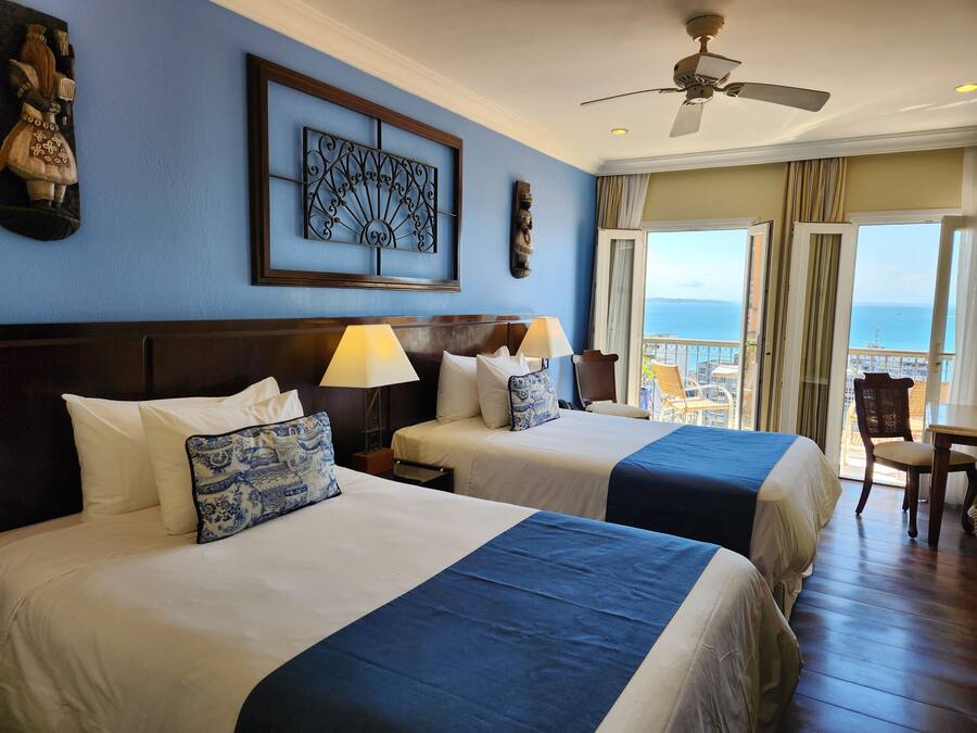 Habitación de hotel con dos camas, ventilador de techo, vista al mar y balcón con mesa y silla.
