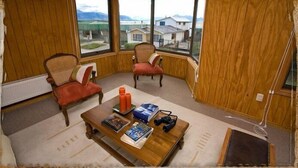 Une pièce aux murs lambrés, avec deux fauteuils, une petite table sur laquelle sont posés des magazines et un appareil photo, et de grandes fenêtres offrant une vue sur des maisons et des montagnes.