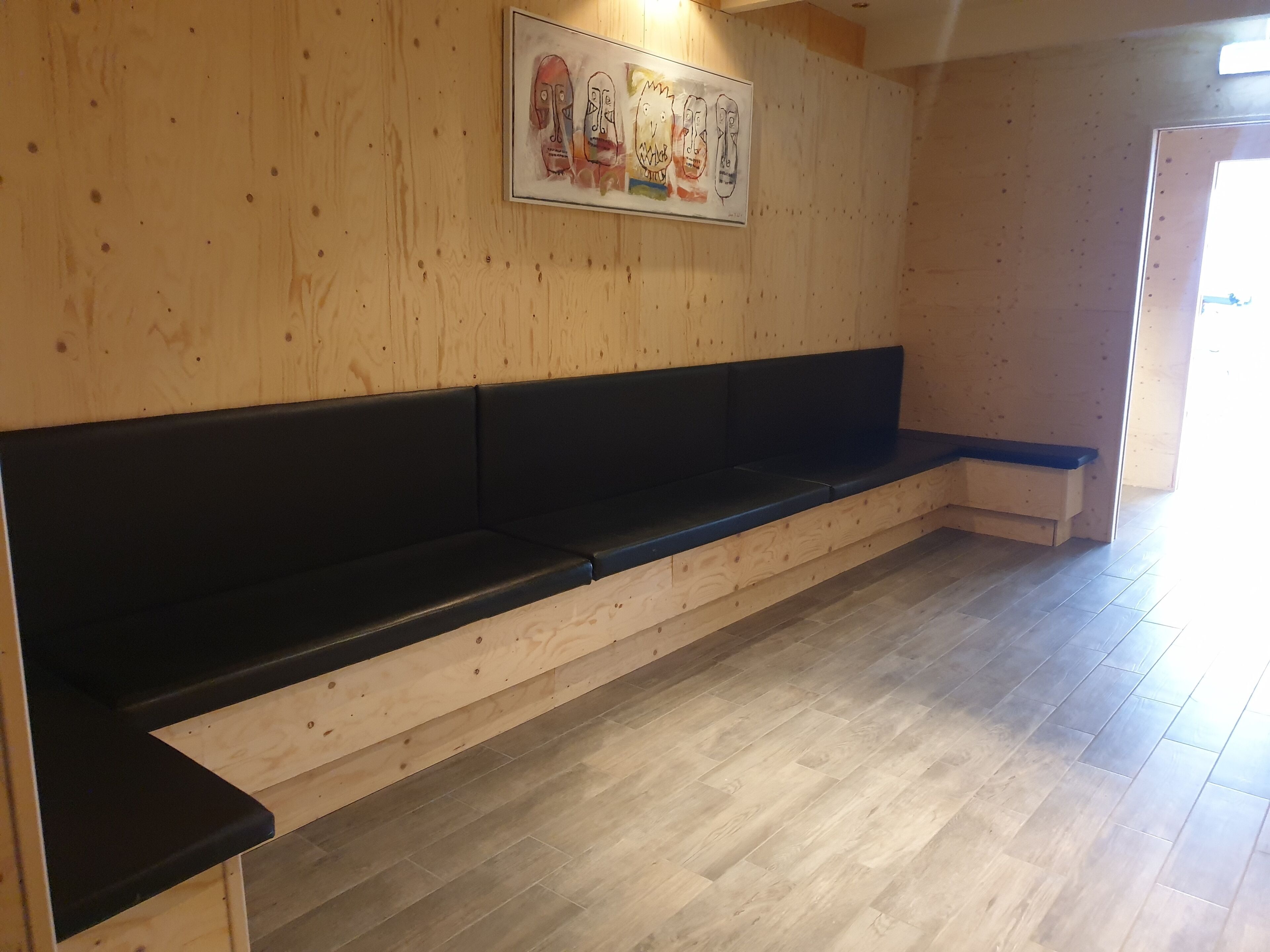 Eine hölzerne Bank mit schwarzen Kissen in einem Raum mit Holzwänden und Fliesenboden.