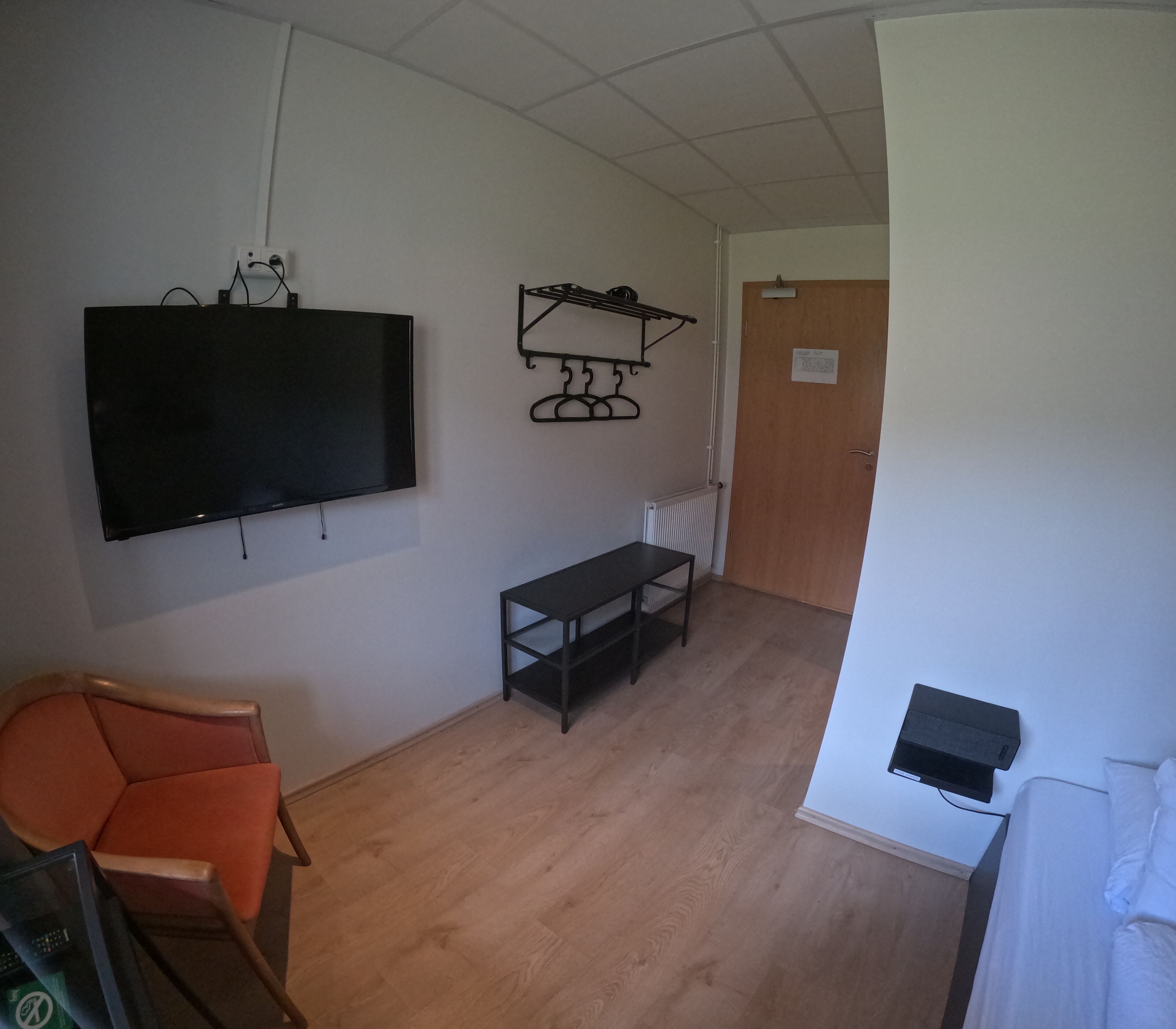 A room with a flat-screen TV mounted on the wall, a wooden floor, a small table with chairs, a wooden door, and a white wall.