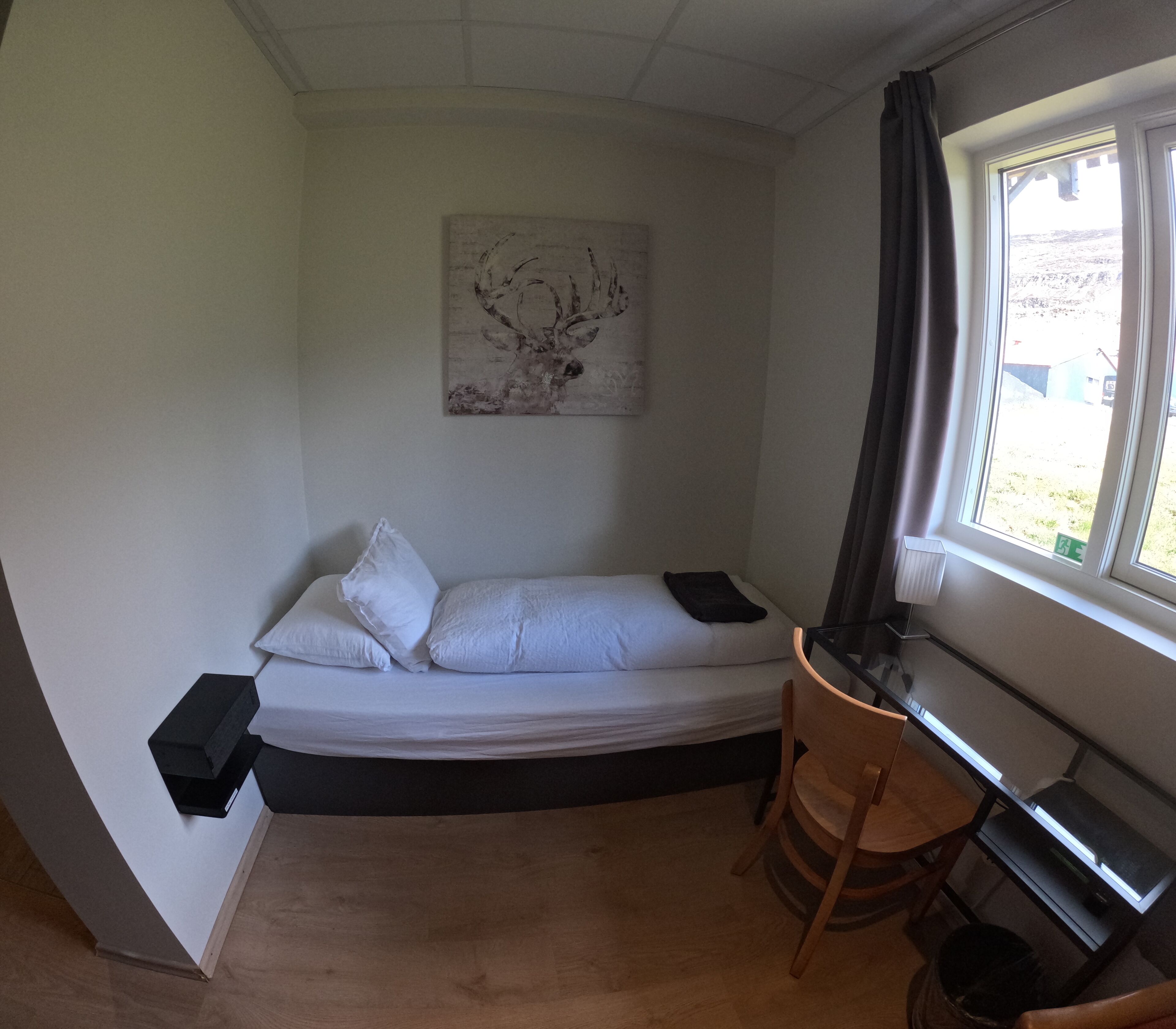 A small, compact room with a single bed, a desk, and a chair. There is a window with curtains, and a painting on the wall.