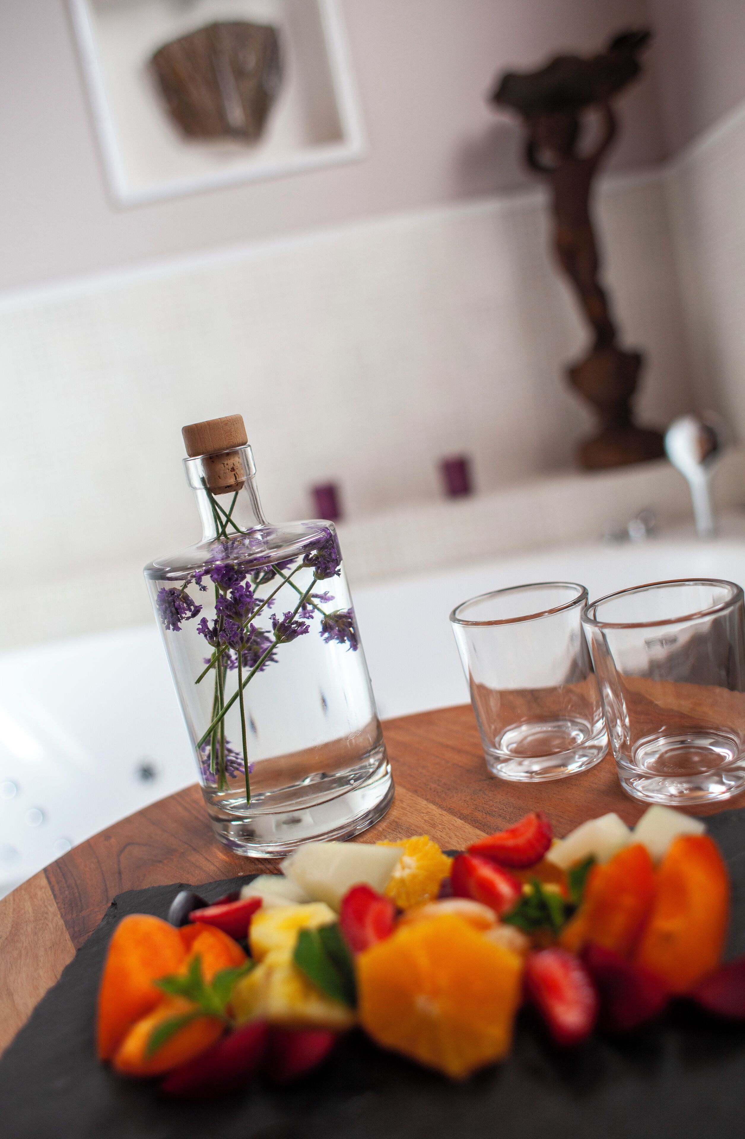 Una botella de licor con tapón de corcho, dos vasos vacíos y una bandeja de frutas sobre una mesa de madera.