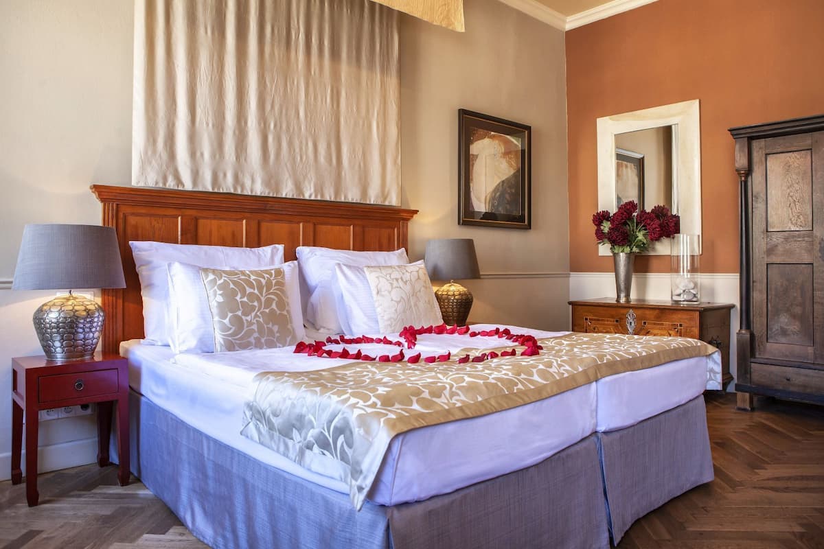 A double bed with white linens, a wooden headboard, and a decorative garland of red flowers. There are two bedside tables with lamps, a mirror, and a vase with red flowers on a dresser.