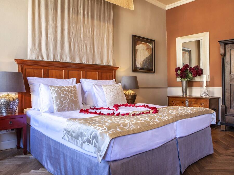 Una cama doble con ropa blanca, cabecera de madera y una guirnalda decorativa de flores rojas. Hay dos mesitas de noche con lámparas, un espejo y un florero con flores rojas sobre un tocador.