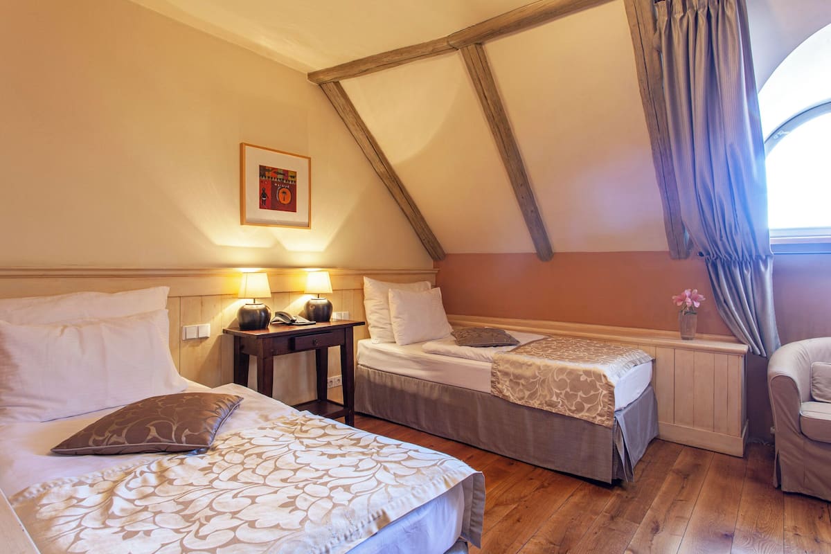 A hotel room with two beds, a wooden ceiling, a nightstand with a lamp, and a window with curtains.