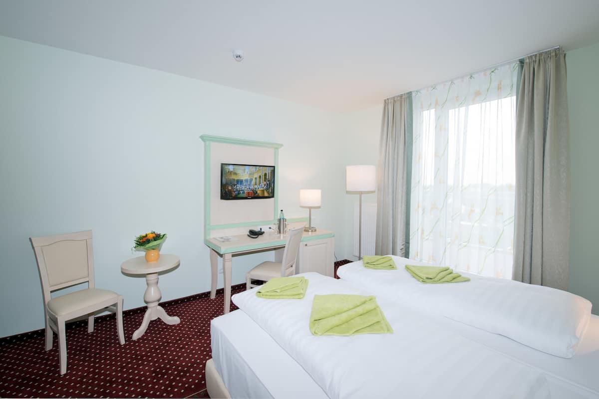 A hotel room with a bed, two chairs, a desk, a small table with a vase of flowers, a painting on the wall, and a window with curtains.