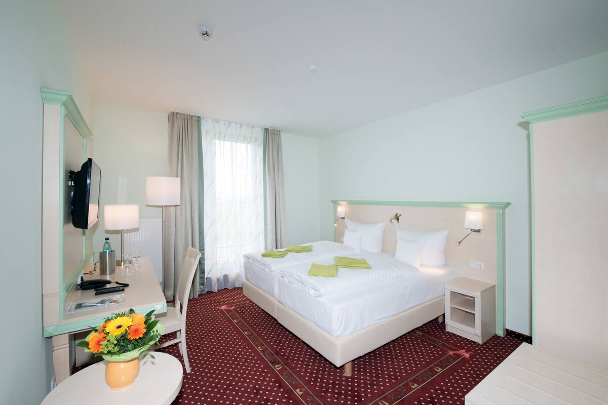 A hotel room with a bed, bedside table, lamp, and a desk with a telephone and a vase of flowers.