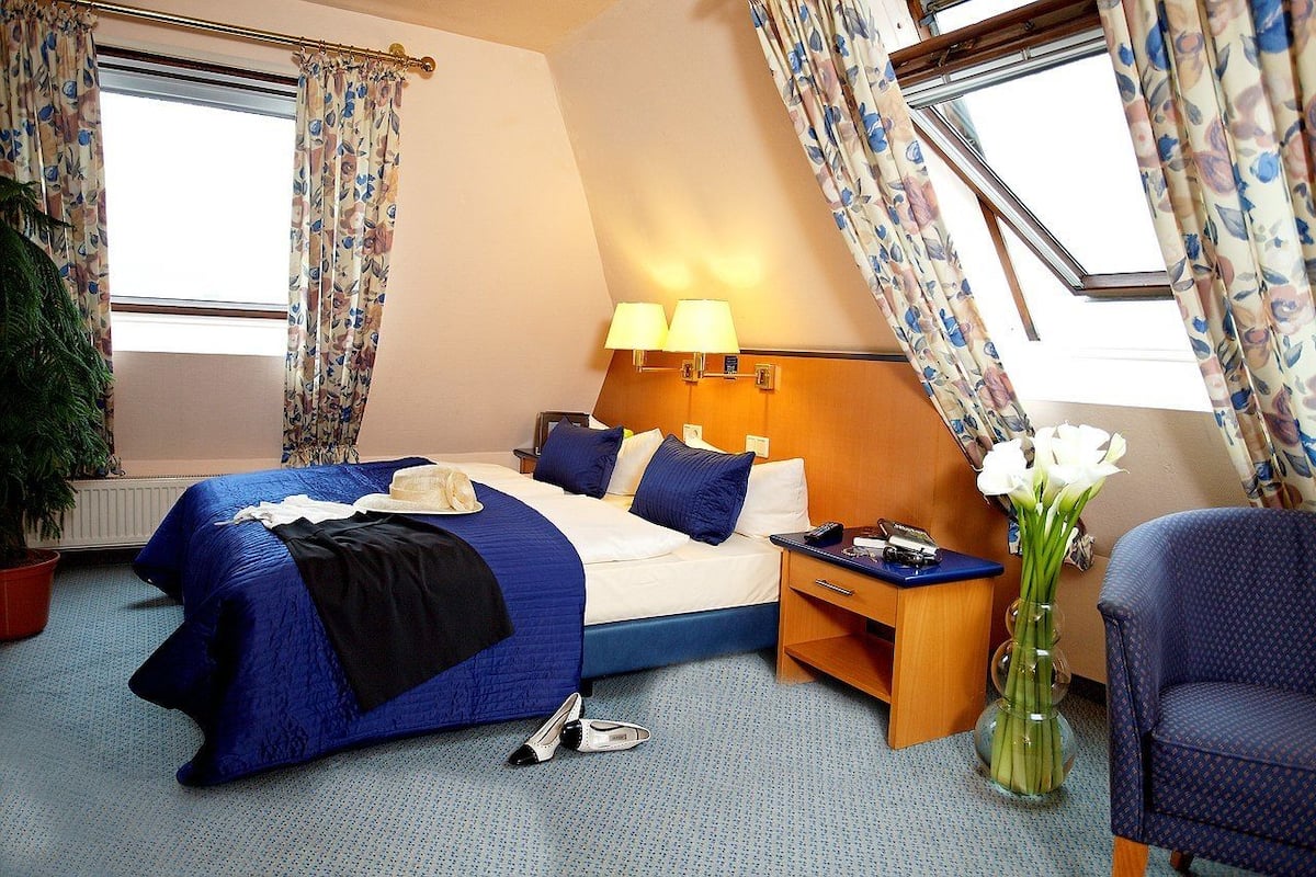 Ein Hotelzimmer mit einem Bett, zwei Nachttischlampen, einem Nachttisch, einem Sessel, einer Topfpflanze und einem Fenster mit Vorhängen.