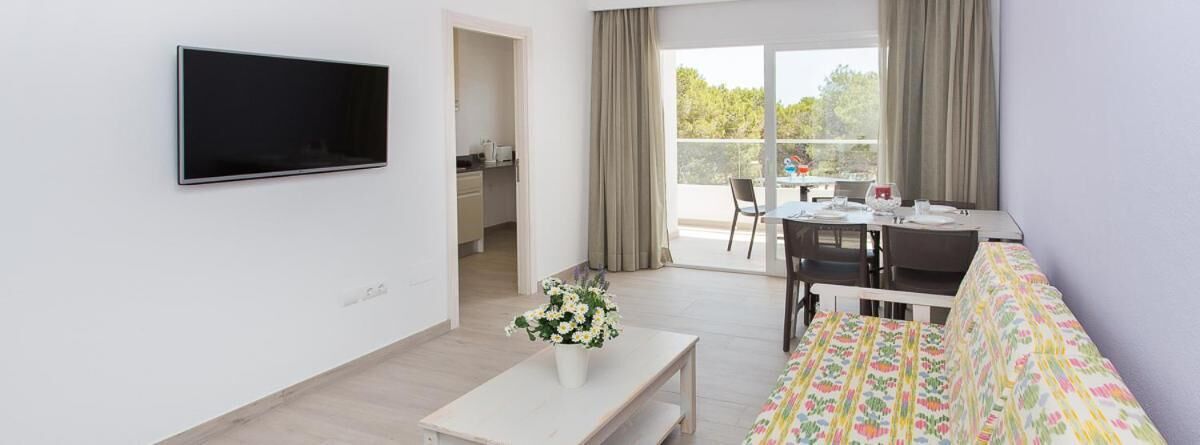 A modern living room with a flat-screen TV, a sofa, a dining table with chairs, and a kitchen area visible through an open door.