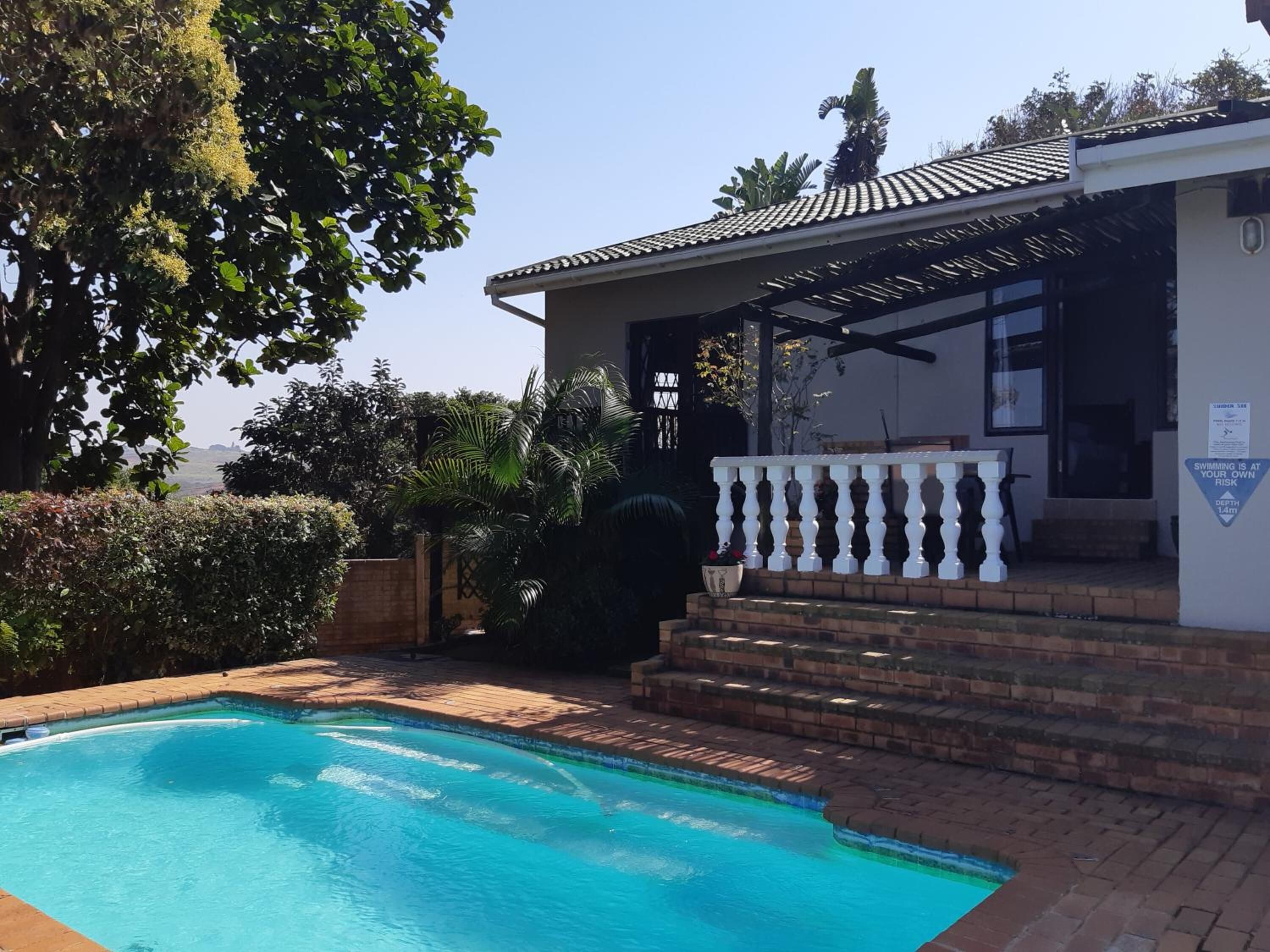 A swimming pool with a covered patio area, surrounded by lush greenery and a clear sky.