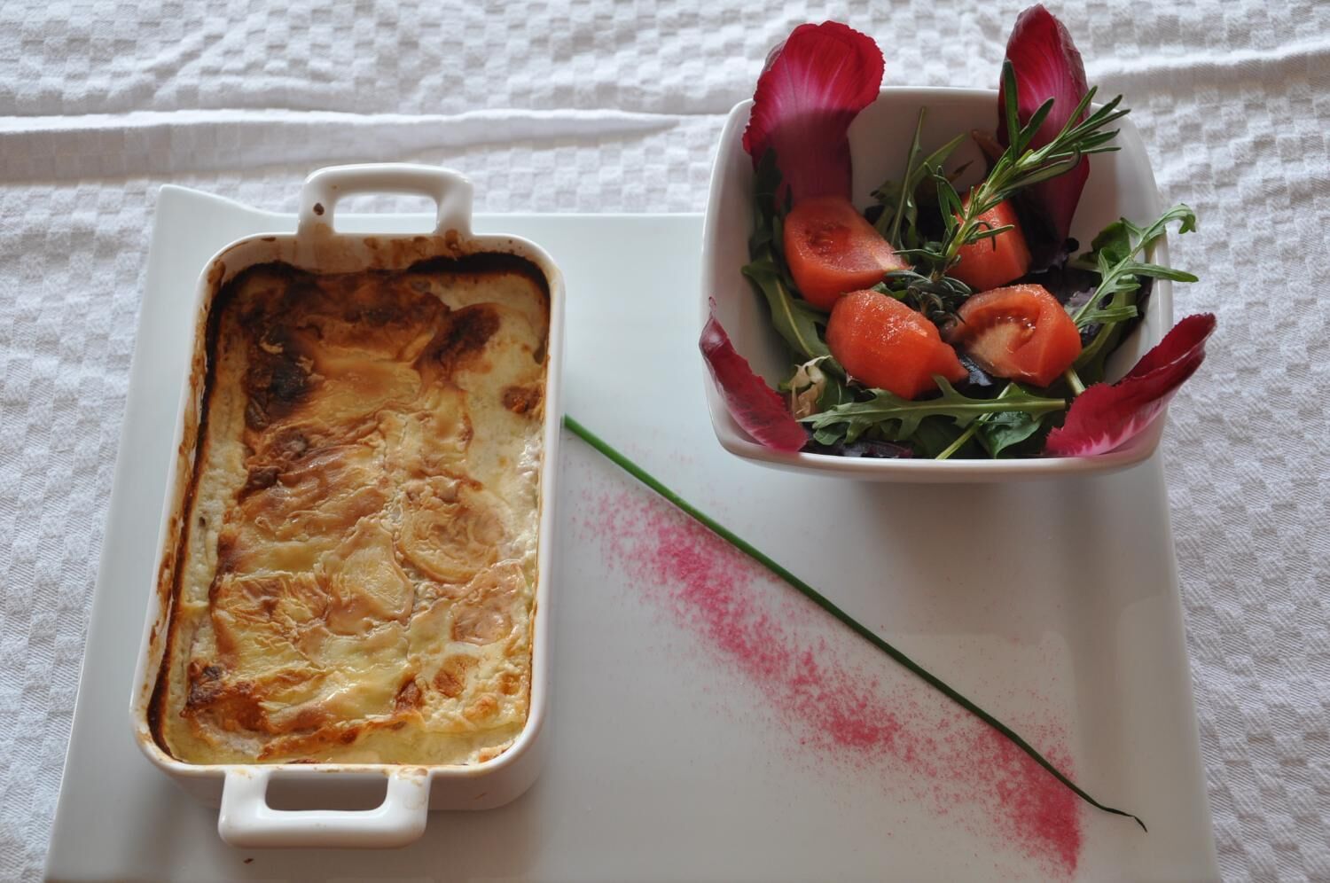 A white rectangular baking dish with a golden-brown top, next to a white bowl with a red flower and green leaves.