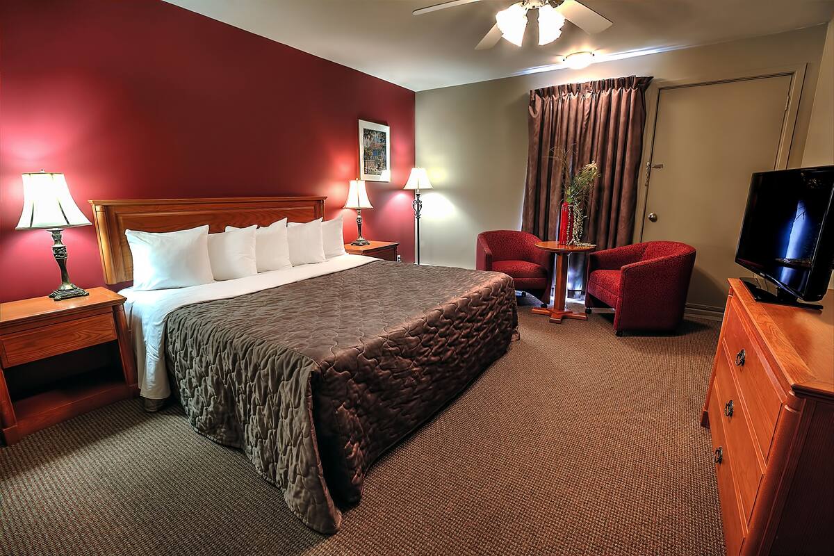 A hotel room with a large bed, two bedside lamps, a TV, a red armchair, a small table, and a wooden dresser.