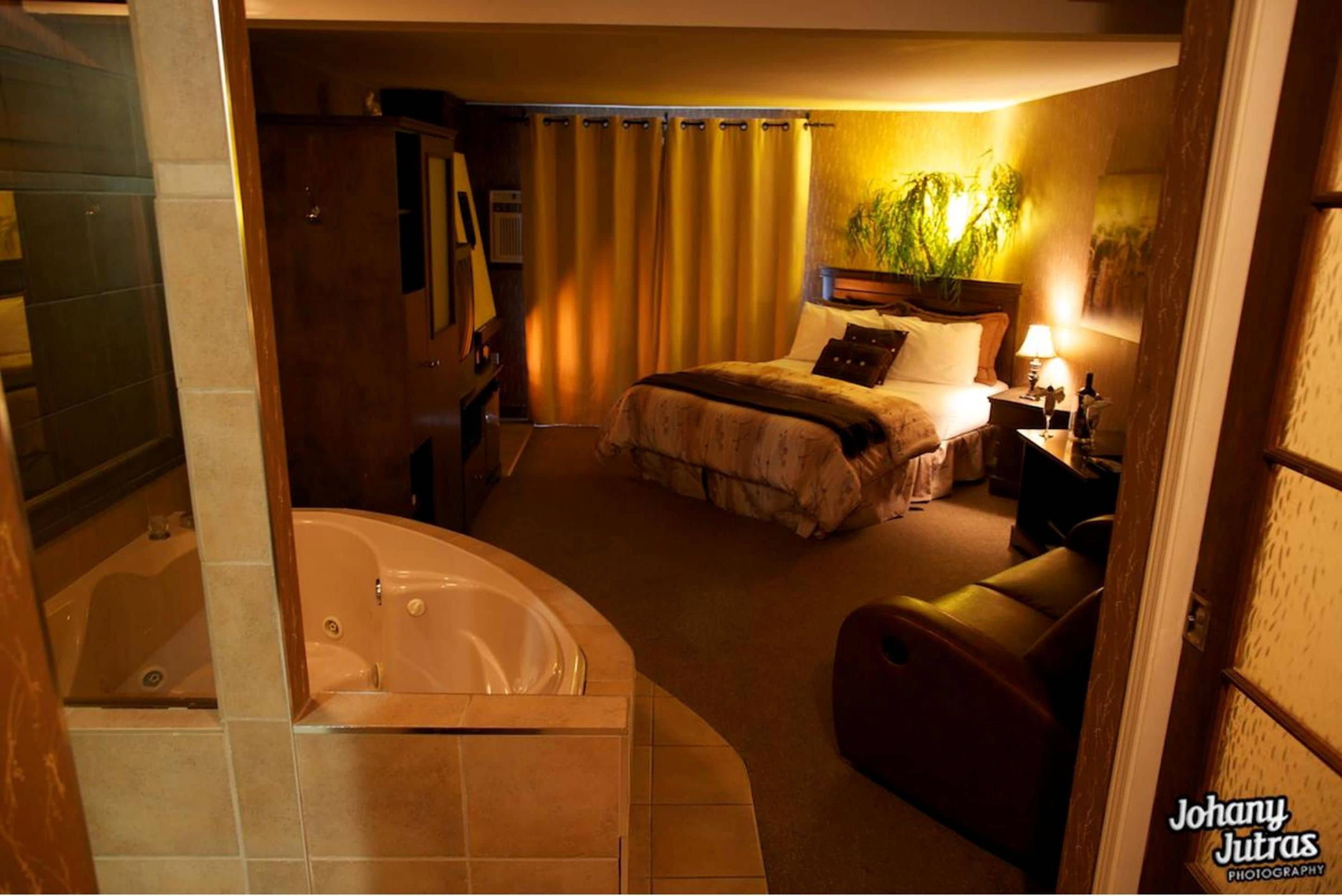 A hotel room with a large bathtub, a bed with a headboard, a nightstand, and a potted plant.