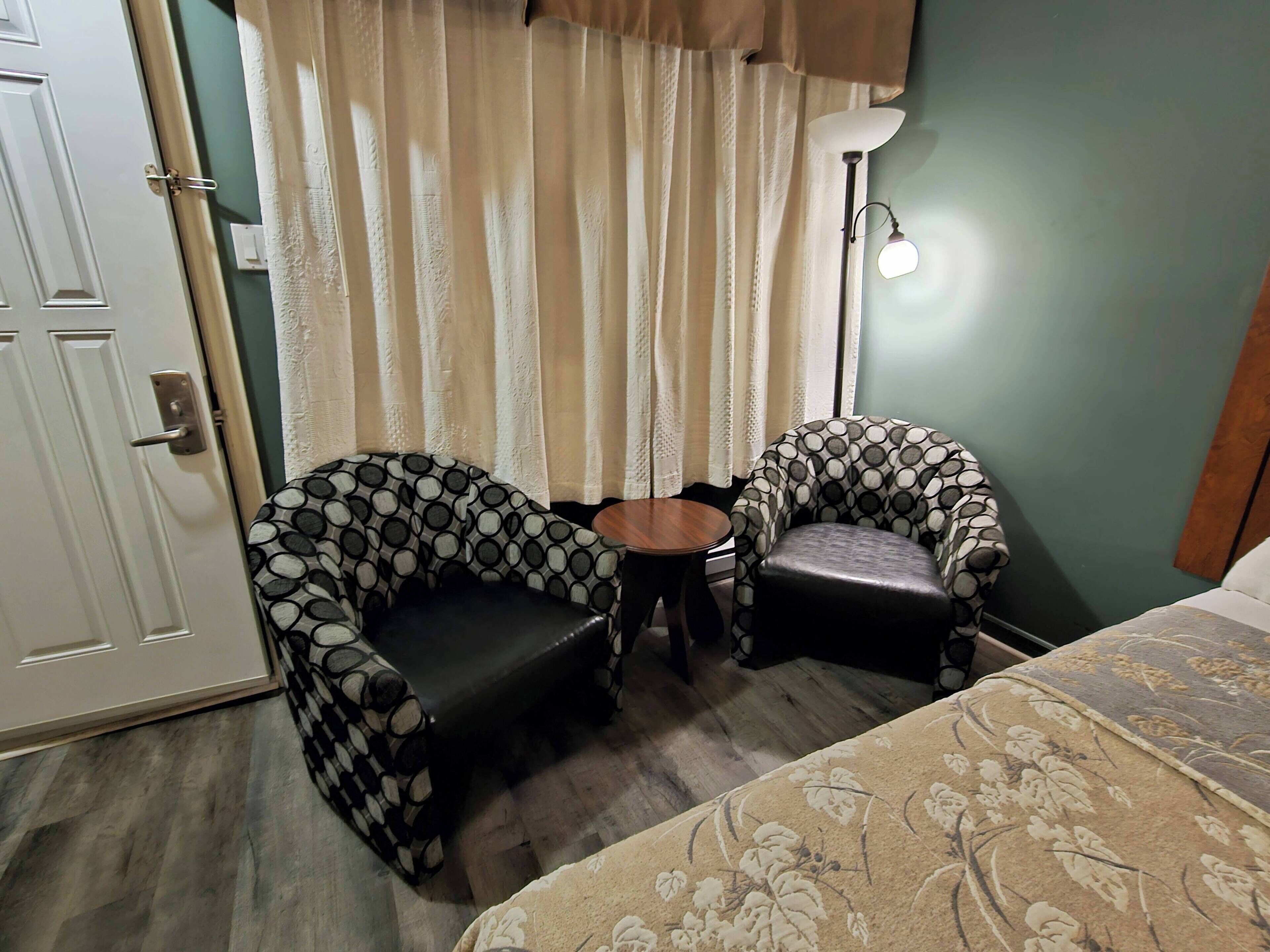 Una habitación con dos sillones estampados, una mesita redonda, una cama con colcha floral, una lámpara de pie y una puerta.