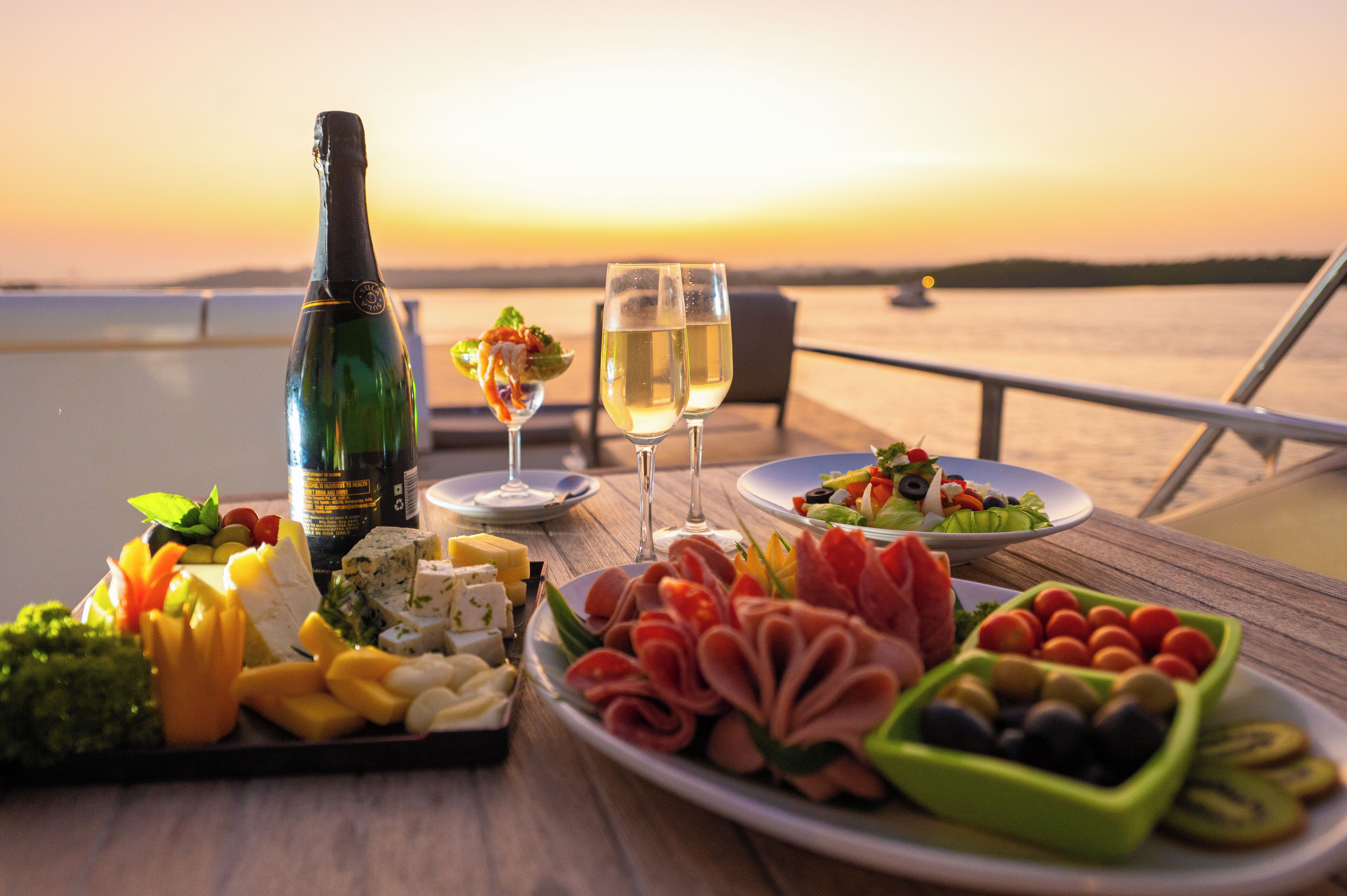 Ein Tisch mit einer Champagnerflasche, zwei Gläsern und verschiedenen Speiseplatten vor einem Sonnenuntergang.