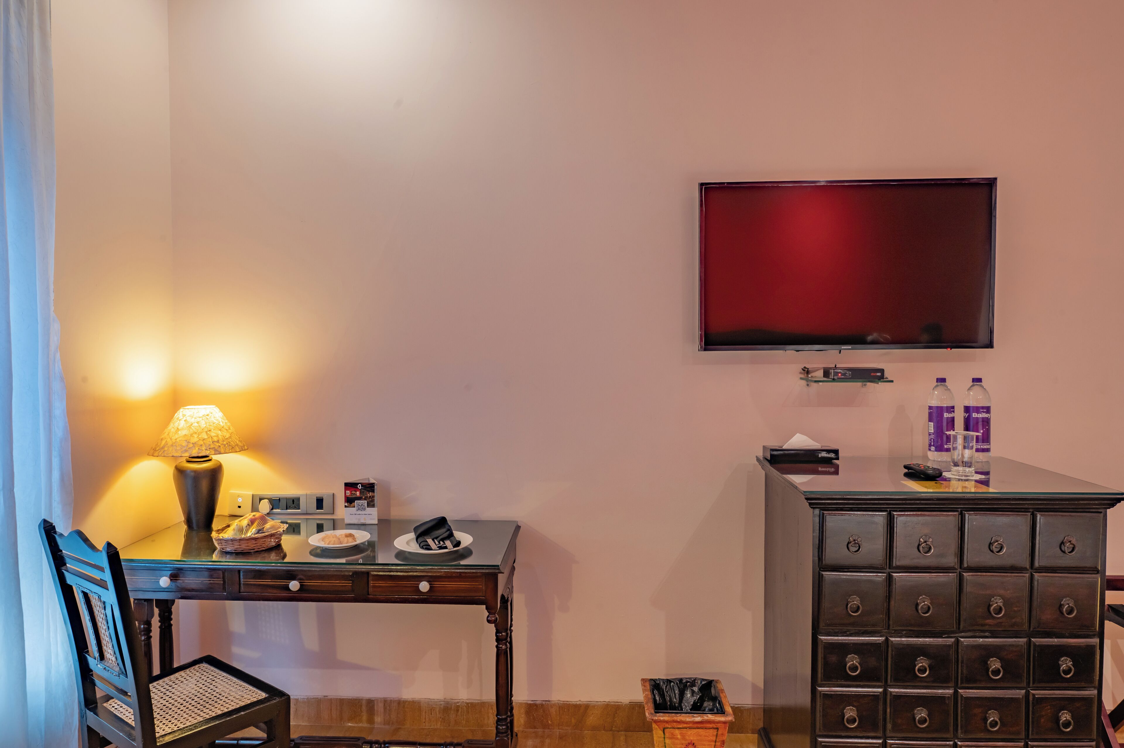 A hotel room with a desk, chair, lamp, television, minibar, and a small table with a basket and plates.