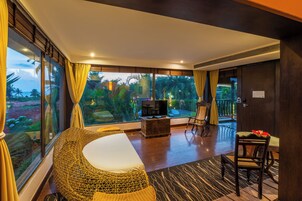 A spacious room with a large bed, a TV, and a dining area with a view of tropical vegetation.