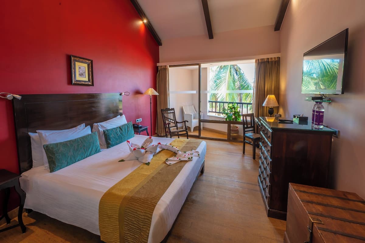 Habitación de hotel con una cama grande, ventilador de techo, televisor, escritorio con lámpara, una silla y vistas a palmeras.