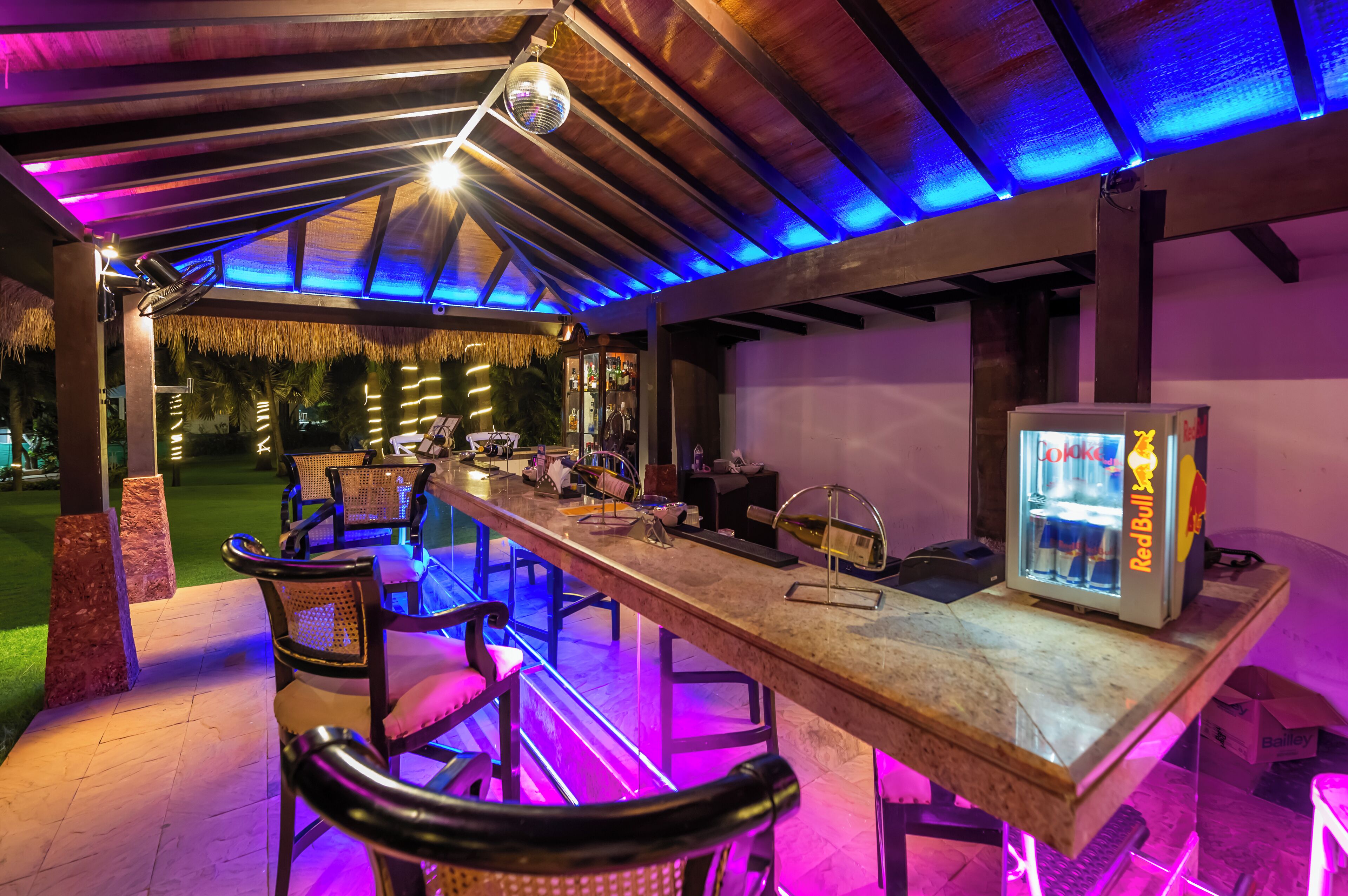 An outdoor bar area with a long wooden table, bar stools, and a Red Bull refrigerator.