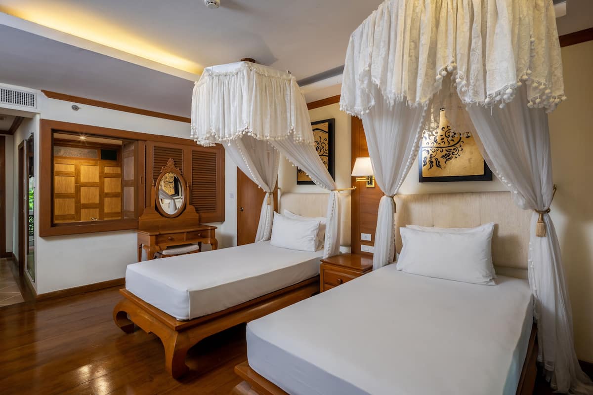 A hotel room with two beds, wooden headboards, a wooden cabinet, a mirror, and a wall-mounted lamp.