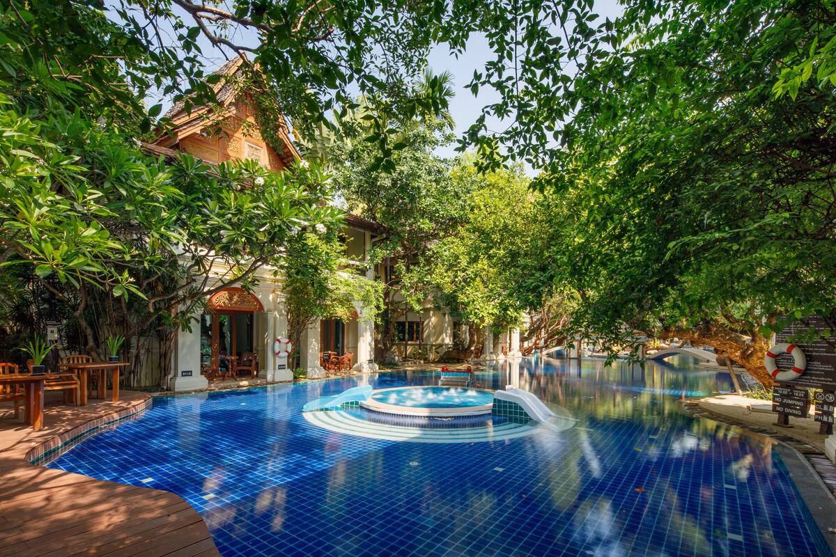 A swimming pool with a circular hot tub, surrounded by lush greenery and a traditional building.