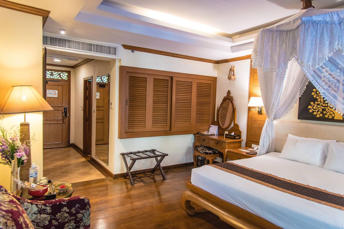 A hotel room with a bed, a desk, a chair, a nightstand, a lamp, a window with shutters, and a decorative mirror.