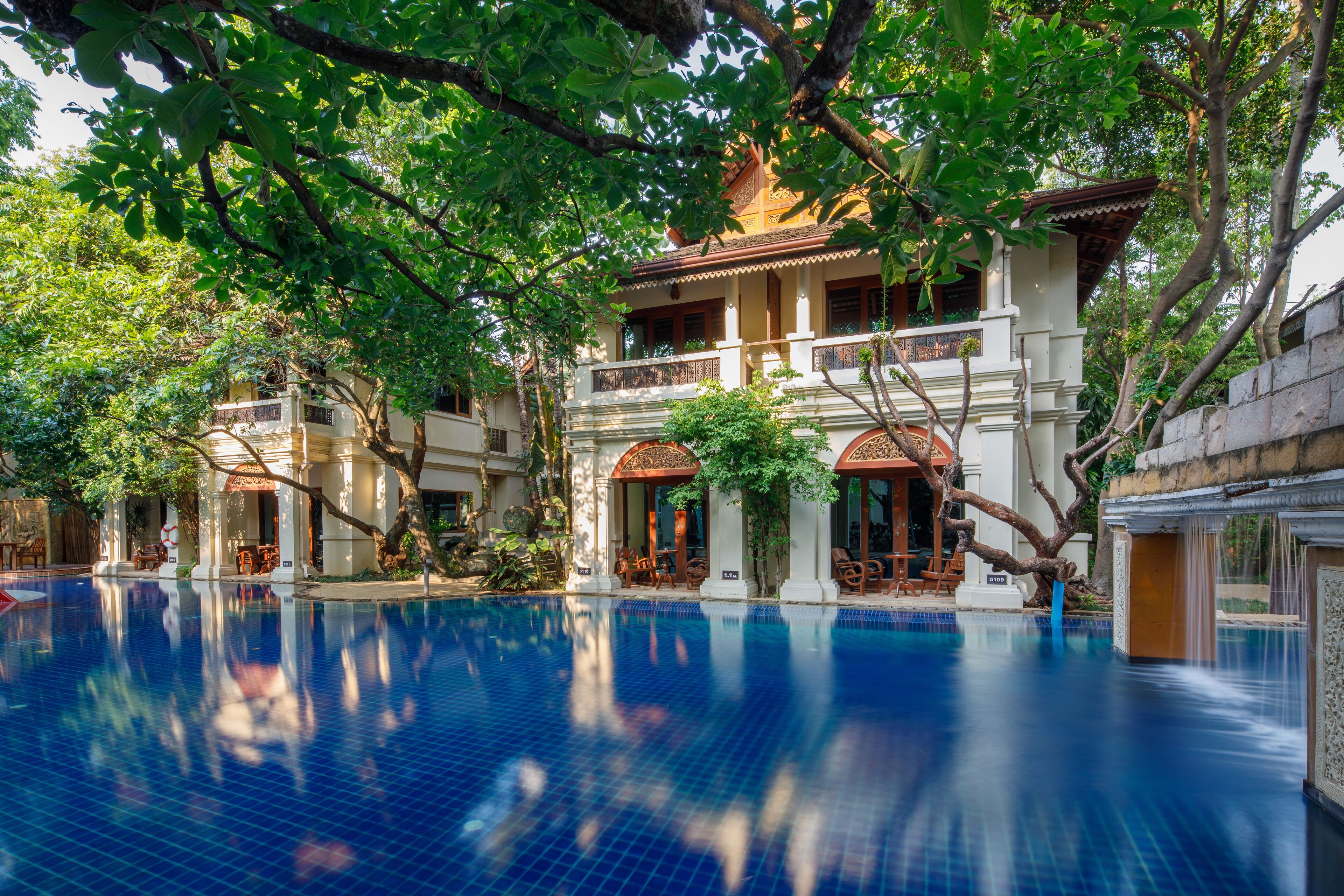A pool area with a clear blue pool, surrounded by trees and a building with a balcony.