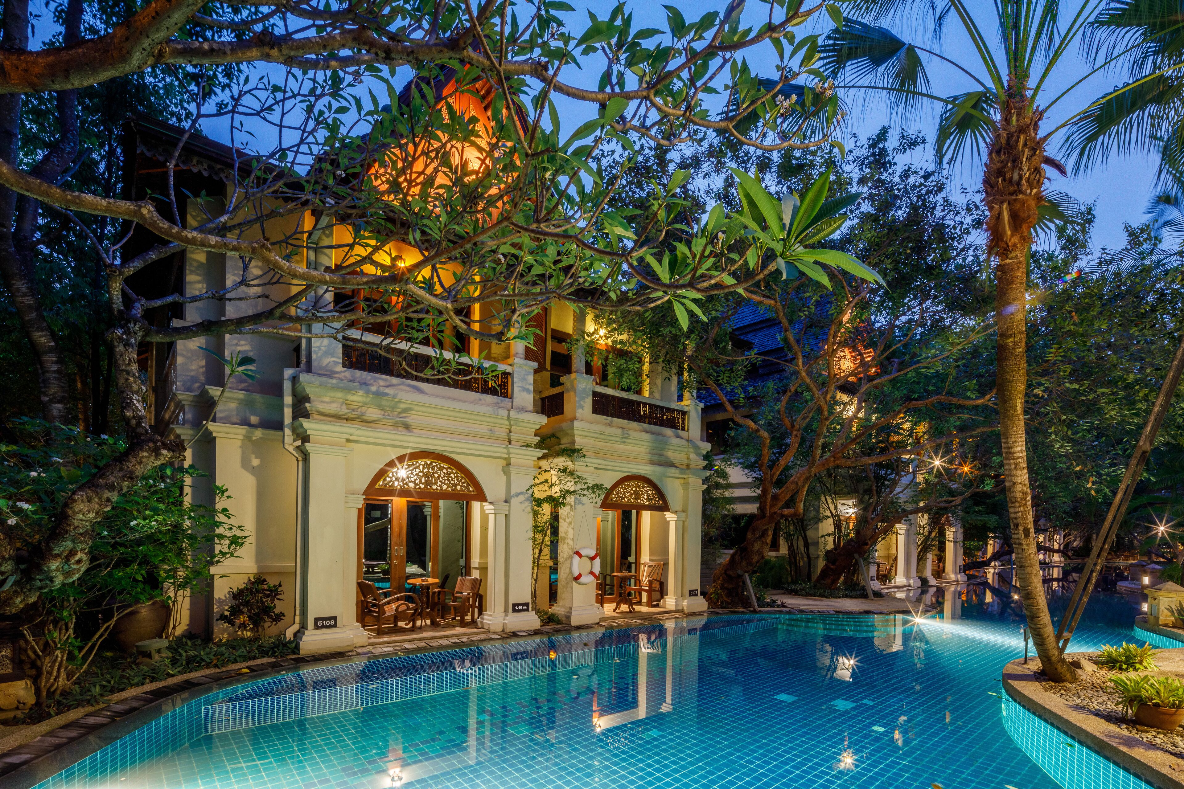 A poolside area with a clear pool, surrounded by lush greenery and a building with arched windows and outdoor seating.