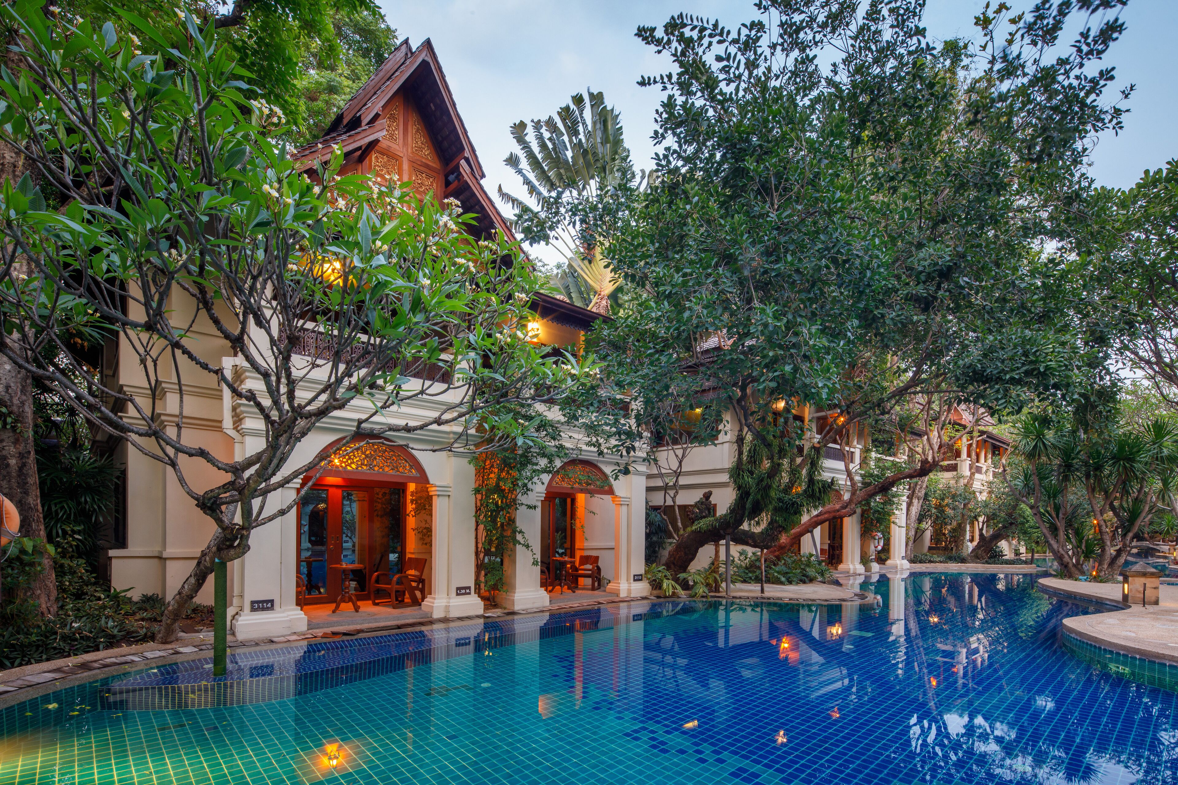 A pool area with a tiled pool, surrounding greenery, and a building with a traditional architectural style.