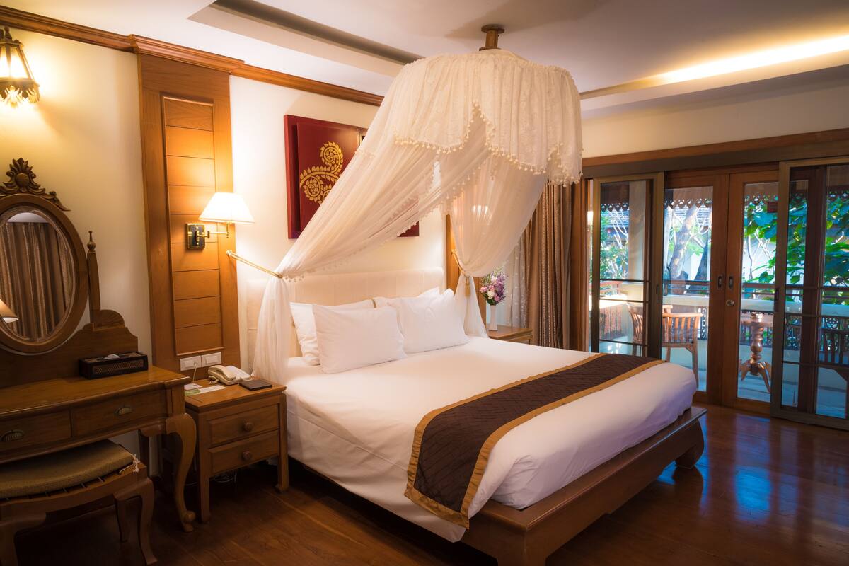 A spacious bedroom with a large bed, wooden furniture, a bedside table with a lamp, and a view through a glass door to an outdoor area.