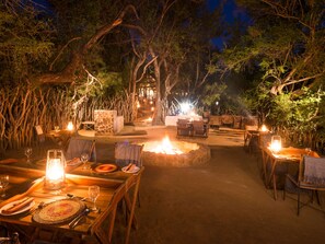 Un área de comedor al aire libre con mesas y sillas dispuestas alrededor de un hogar central, rodeado de árboles.