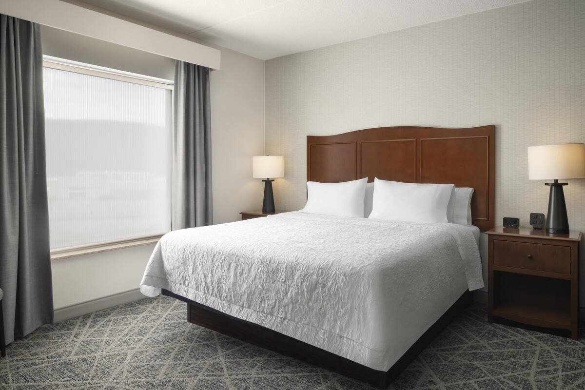 A hotel room with a large bed, two bedside tables with lamps, a window with curtains, and a patterned carpet.