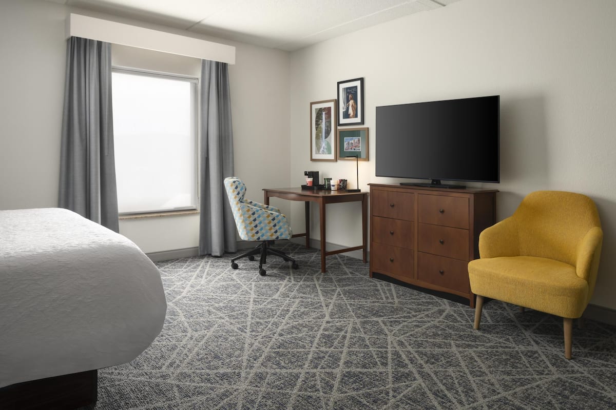 A hotel room with a bed, a desk with a flat-screen TV, a yellow armchair, and a patterned carpet.