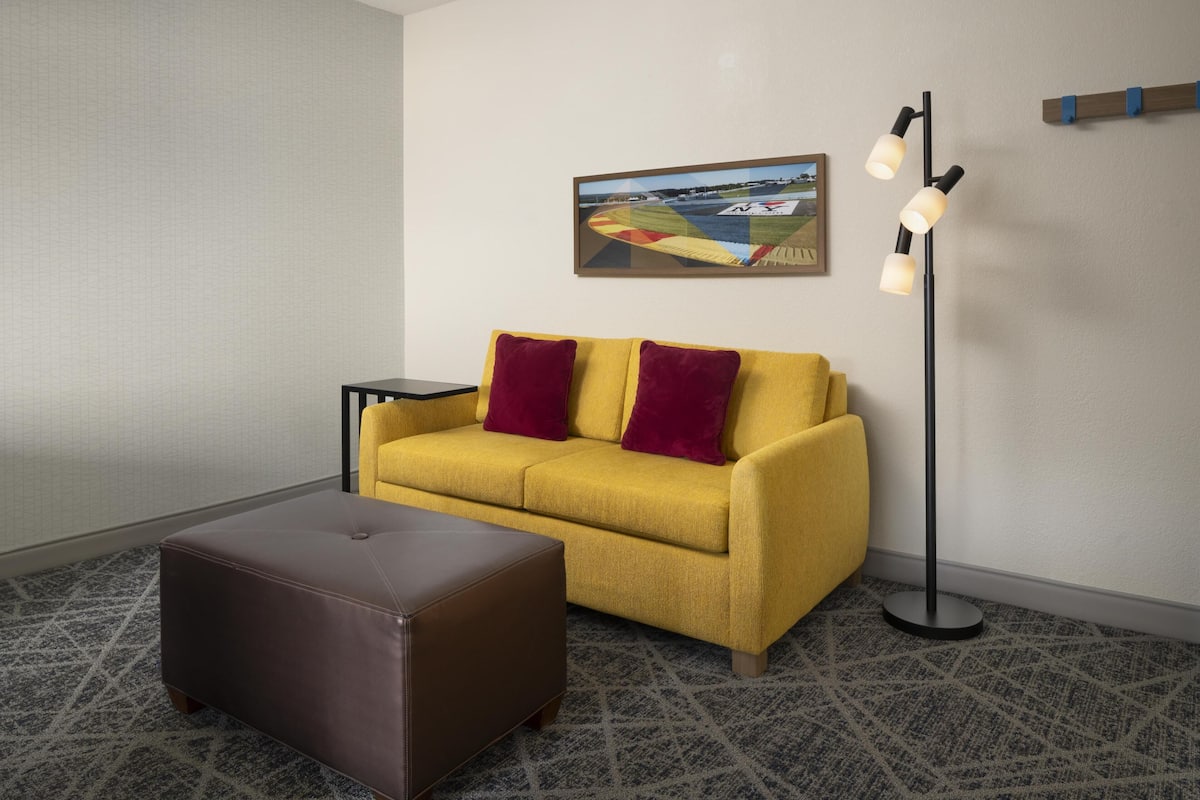A room with a yellow sofa, a brown ottoman, a floor lamp, and a framed picture on the wall.