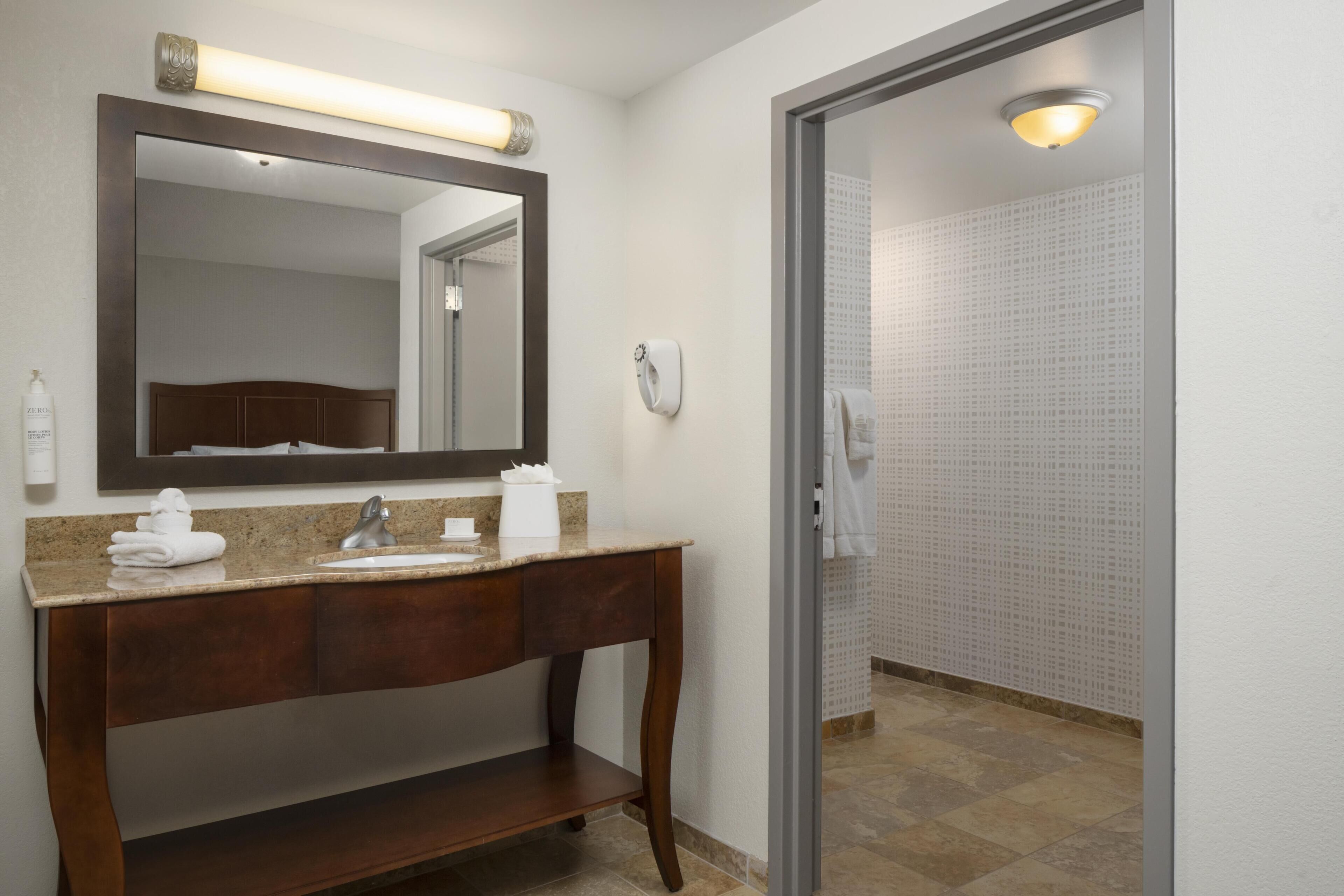 Un baño con tocador de madera, un espejo grande, dispensador de toallas y ducha con pared de azulejos.