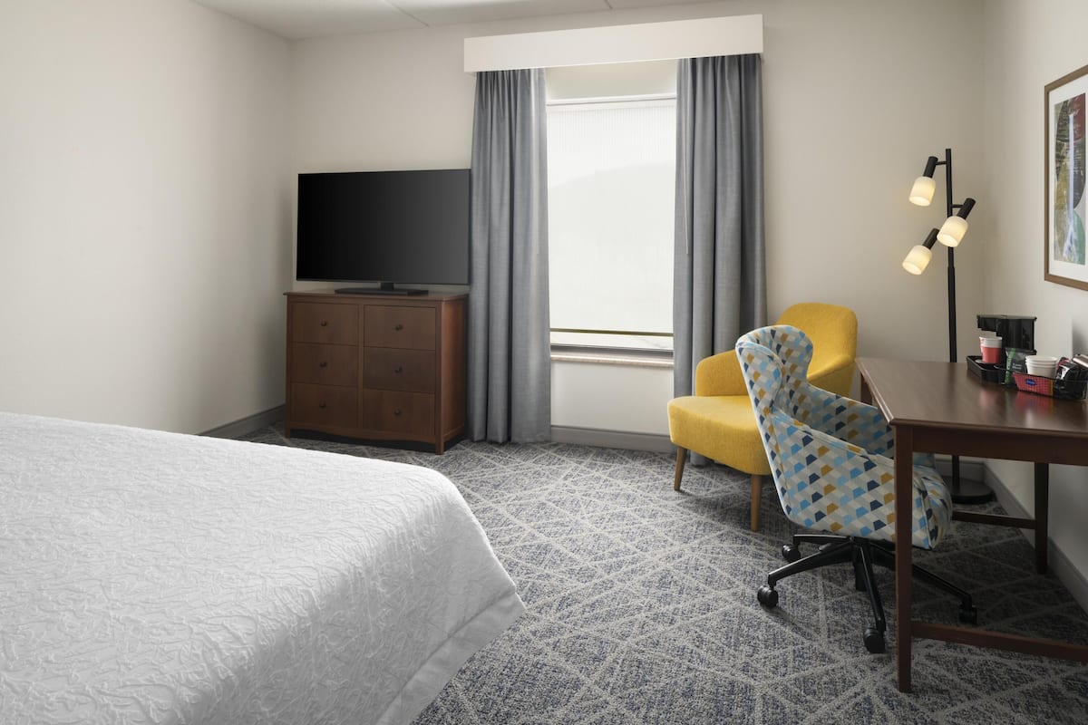 A hotel room with a bed, a TV on a wooden dresser, a yellow chair, a desk with a lamp, and a window with curtains.