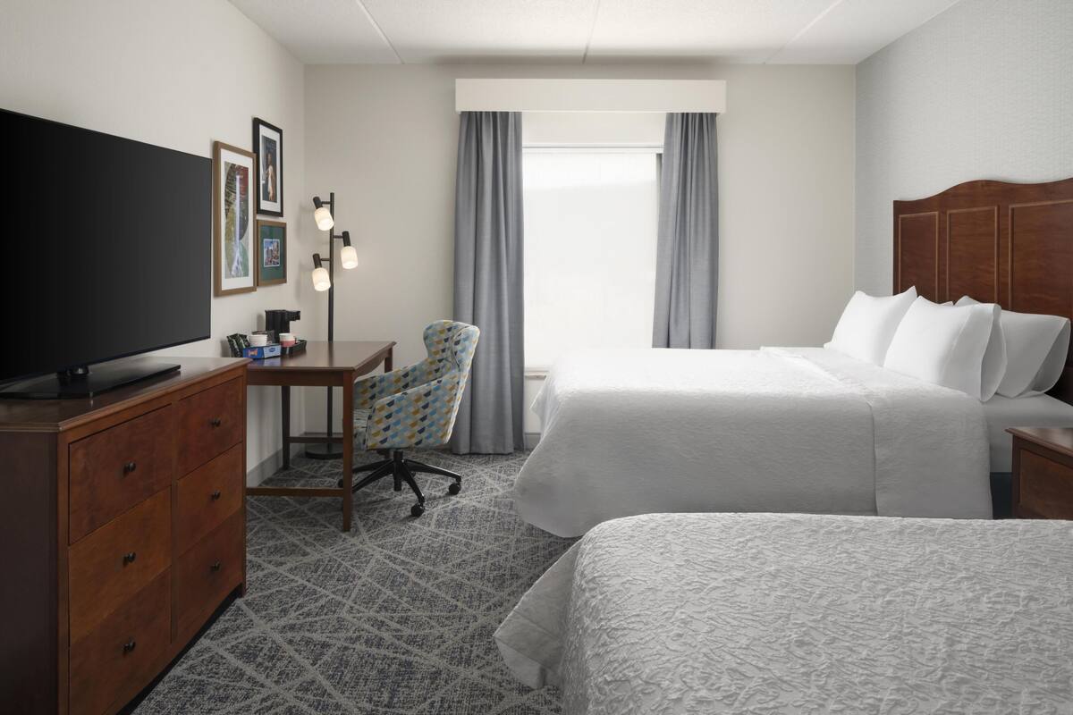 A hotel room with a bed, a desk with a chair, a television, and a window with curtains.
