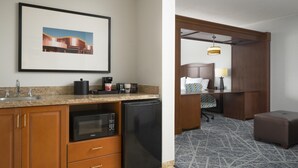 Quarto de hotel com cozinha compacta, uma cama com cabeceira, escrivaninha com cadeira, luminária e tapete estampado.