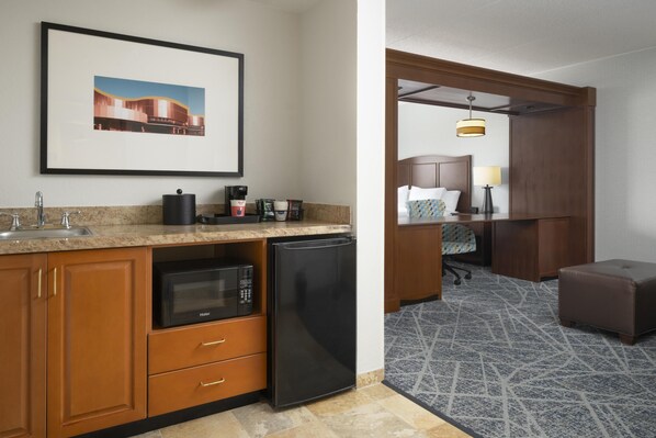 A hotel room with a kitchenette, a bed with a headboard, a desk with a chair, a lamp, and a patterned carpet.