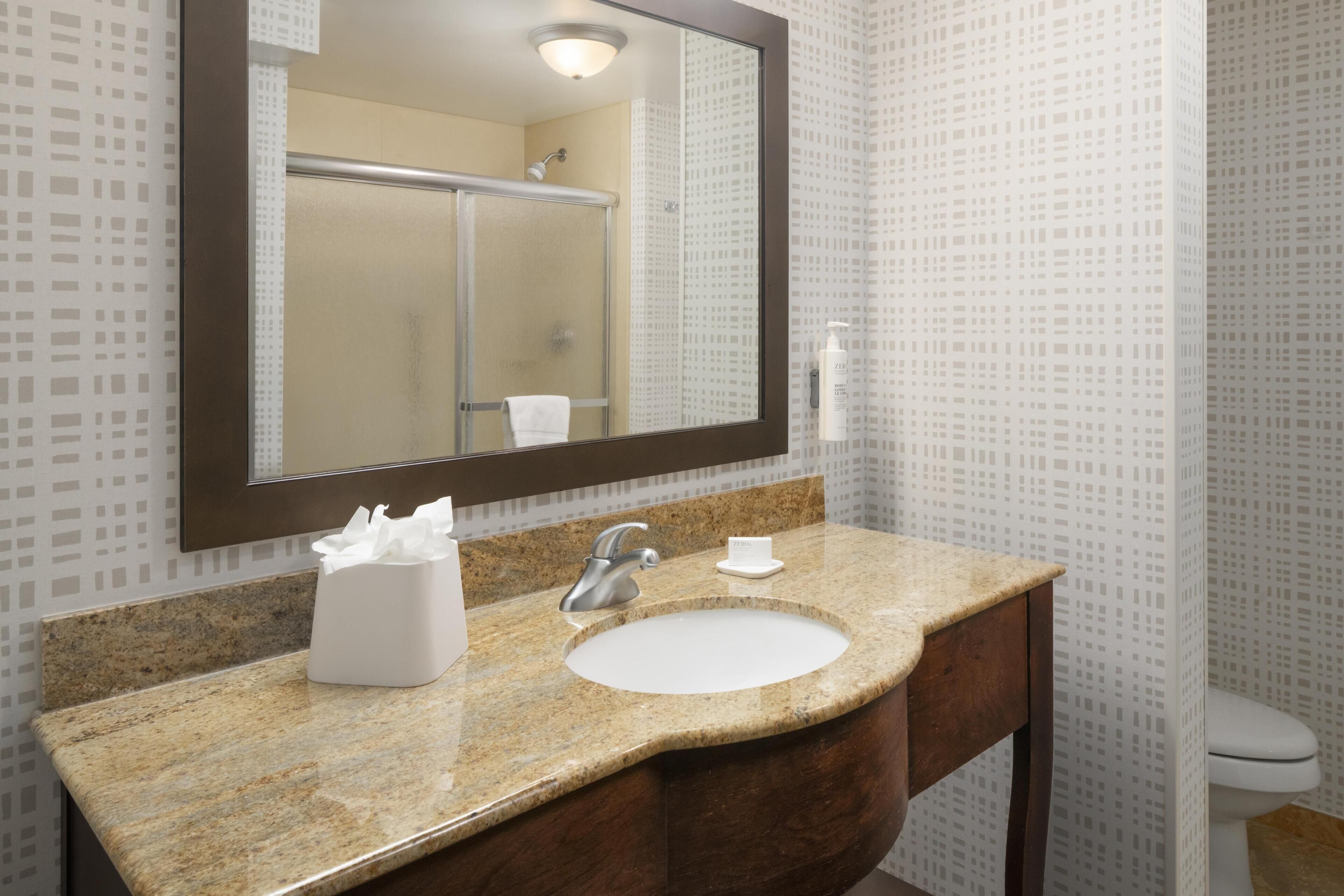 Un baño con lavabo de encimera de granito, espejo, dispensador de toallas y una ducha visible en el reflejo.