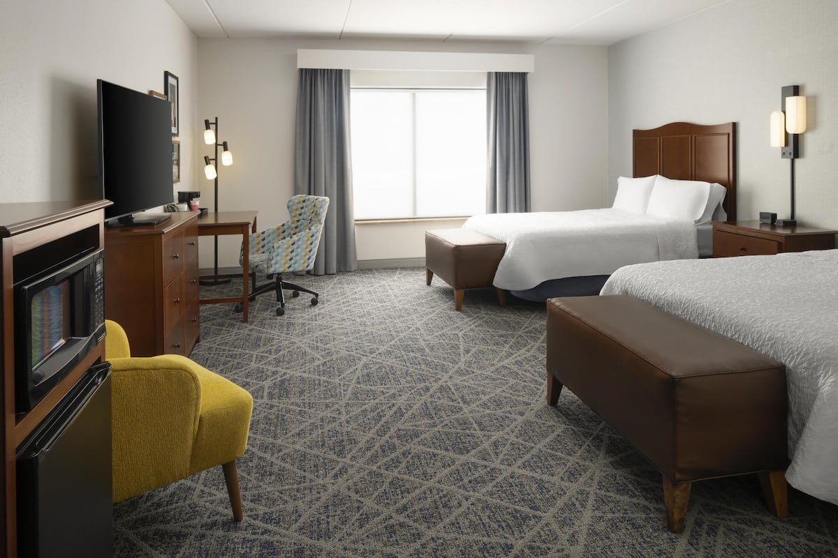 A hotel room with a bed, a desk with a TV, a chair, a bench, and a window with curtains.